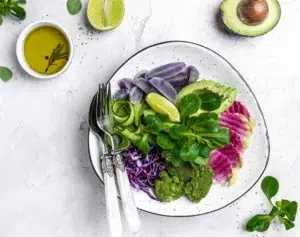 Ein Teller mit Vogerlsalat, Gurke, Broccoli, Rotkraut und Avocado garniert mit einer Scheibe Zitrone. Links am Teller liegen ein Löffel und eine Gabel.
