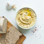 Eine weiße Schale mit Hummus daneben liegen ein Laib Brot, Chiliflocken und mehrere Knoblauchzehen.