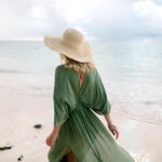 Eine Frau die in der Sonne am Strand spazieren geht. Im Hintergrund sieht man das blaue Meer.