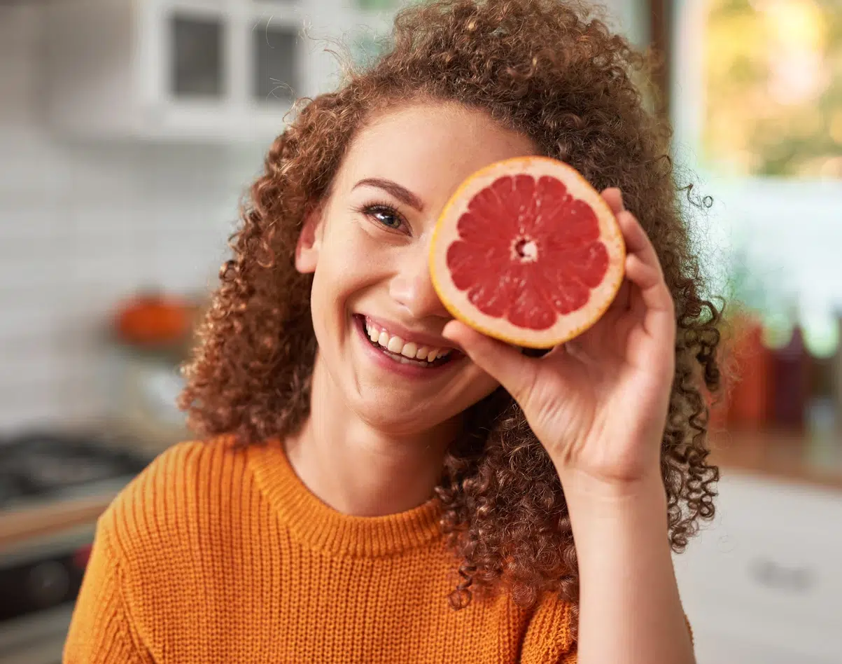 Junge Frau mit roten gelockten Haaren. Sie trägt einen orangen Pullover und hält sich eine halbe Grapefruite vor das linke Auge.