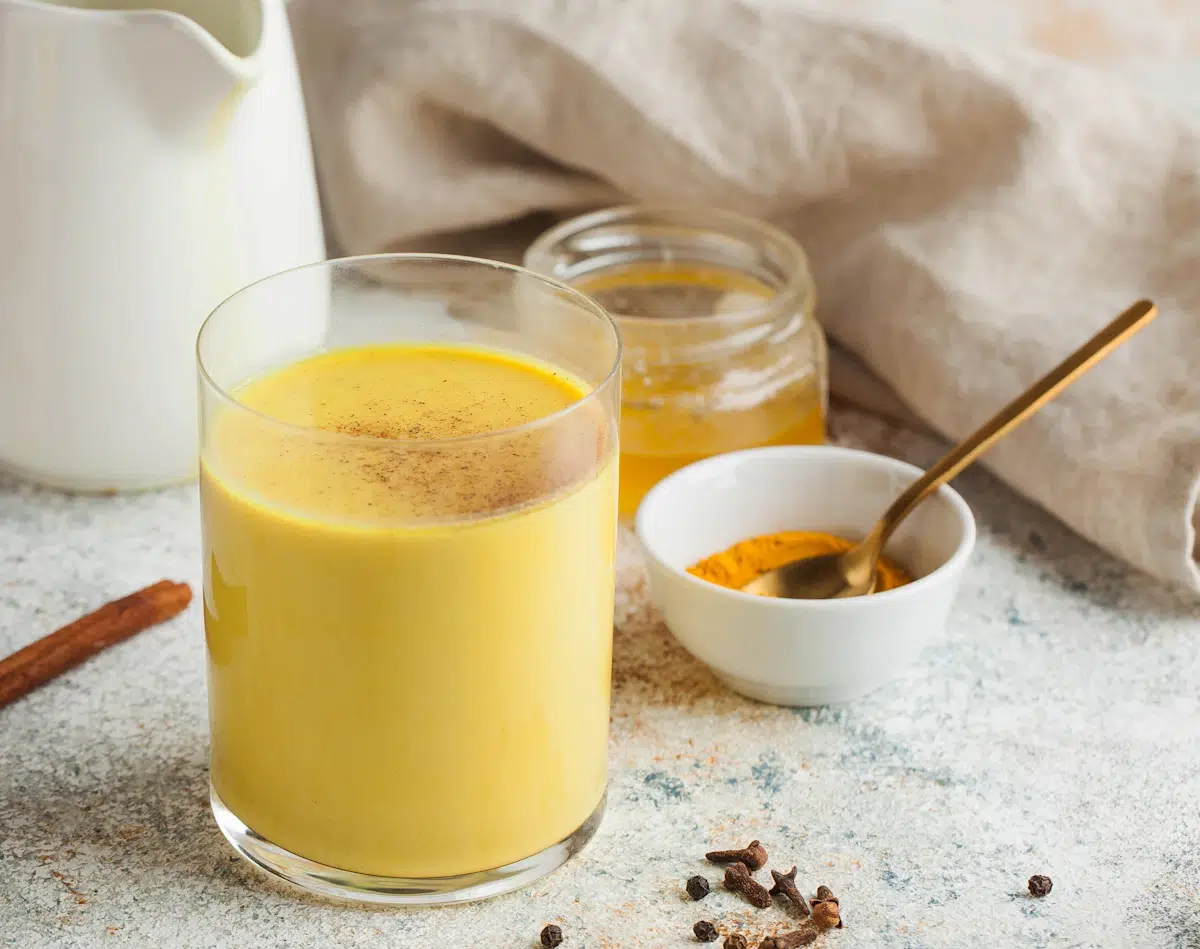 Ein Glas Goldene Milch mit Kurkuma. Danebene steht eine kleine Schüssel mit Curcuma Pulver undein Glas mit Agavendicksaft.