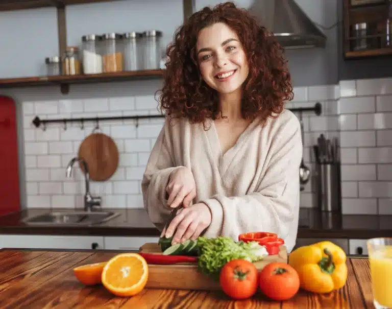 Frau die in der Küche steht und auf einem Holzbrett Gurken in Scheiben schneidet. Daneben liegen noch Paprika, Orangen und Tomaten.